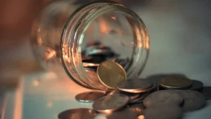 عملات معدنية تنسكب من برطمان زجاجي في ضوء دافئ ترمز إلى اختيار مكان آمن لمدخرات الطوارئ