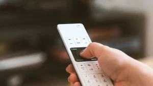 يد تمسك جهاز تحكم عن بعد أمام شاشة في غرفة معيشة دافئة ترمز إلى اشتراكات البث المنزلية