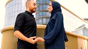 زوجان عربيان، رجل بقميص أسود وامرأة بحجاب كحلي وعباءة مطرزة، يمسكان بأيدي بعضهما أمام مبنى حديث بواجهات ذهبية في رحلة شهر العسل